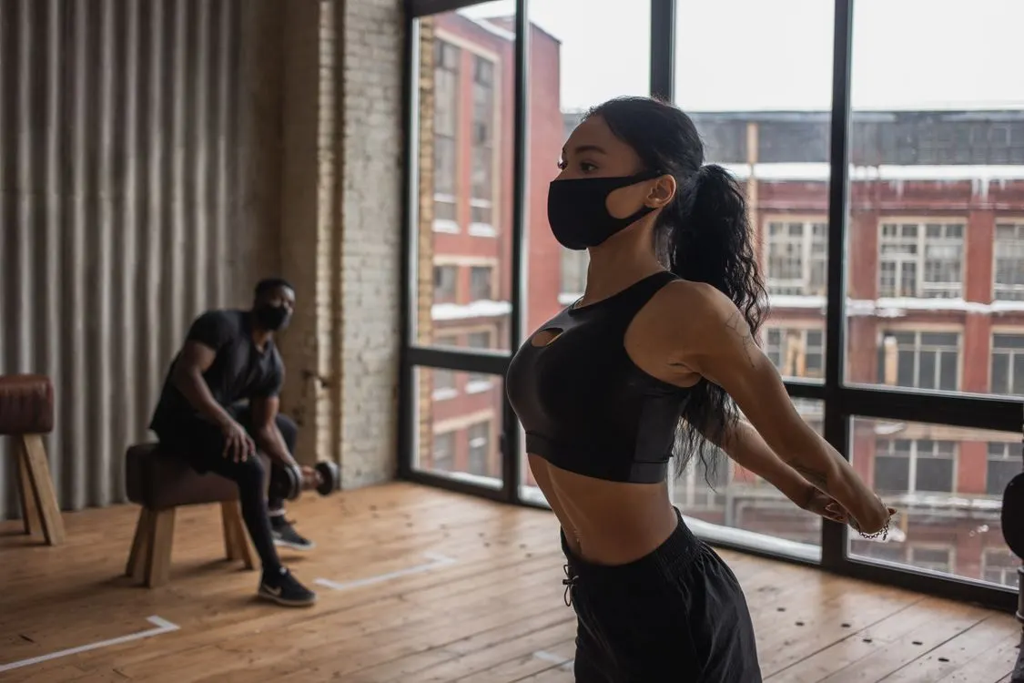 Fit African American sportswoman in mask stretching arms while training in gym during coronavirus pandemic with man lifting dumbbell